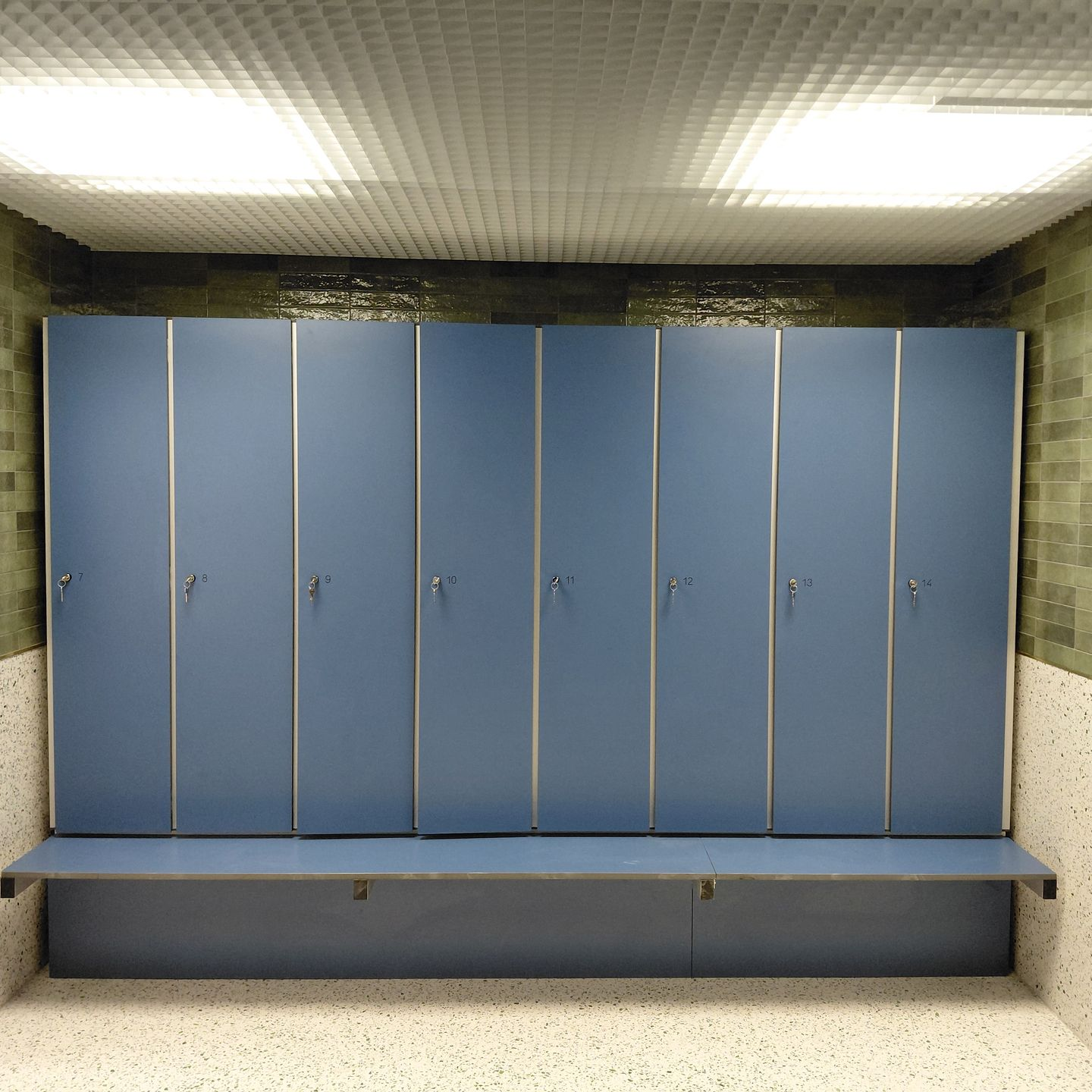 Vestiaire vide avec casiers gris, bancs et murs et sol blancs.