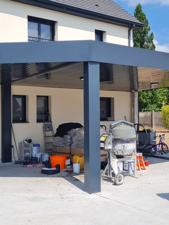 Chantier de construction sous un abri d'auto gris ; des outils, une bétonnière et des seaux de peinture sont visibles.