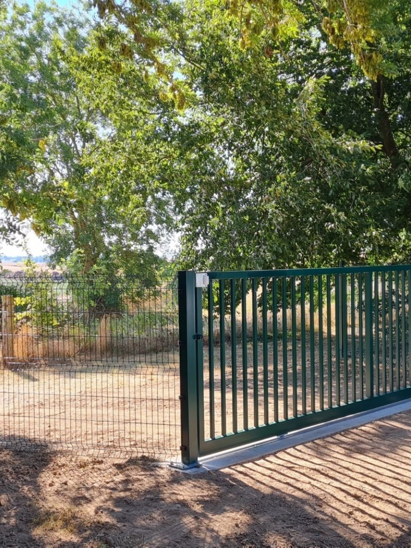 Portail métallique vert, partiellement ouvert, sur un chemin de terre, avec des arbres en arrière-plan.