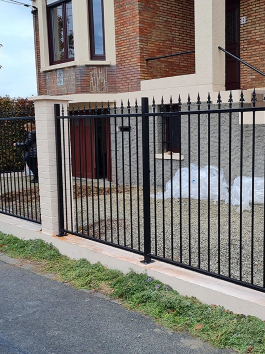 Clôture métallique noire devant un bâtiment en briques. Une personne se tient derrière, l'herbe au premier plan.