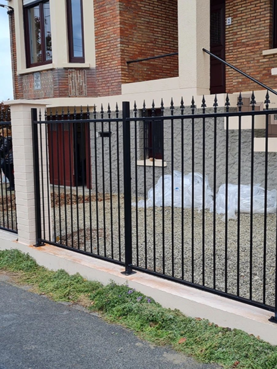 Clôture en métal noir devant une maison en briques aux accents beiges, le long d'une route avec de l'herbe.