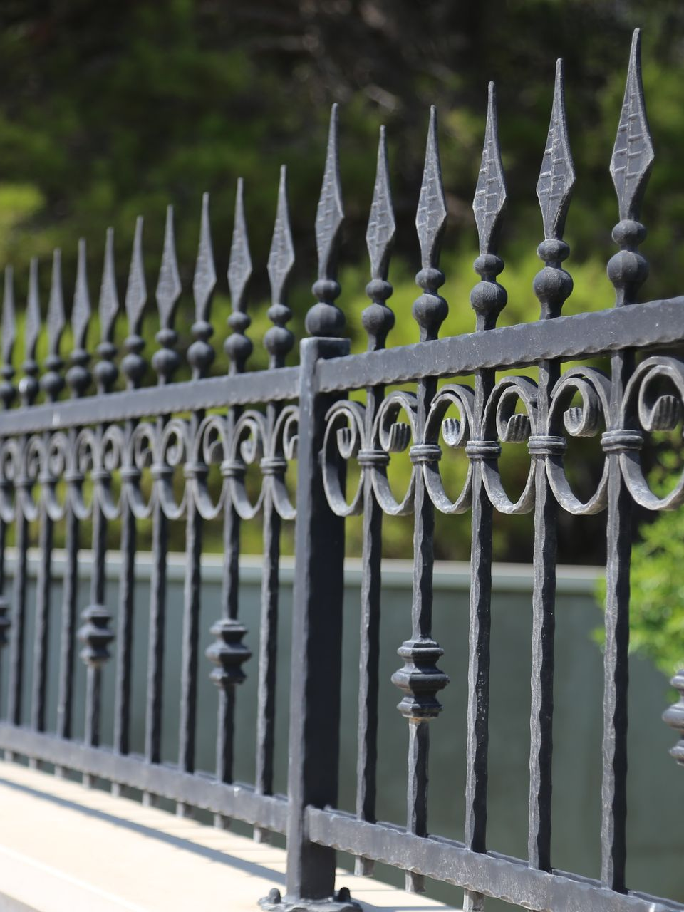 Clôture en fer forgé noir avec pointes décoratives, le long d'un mur en pierre claire à fond vert.