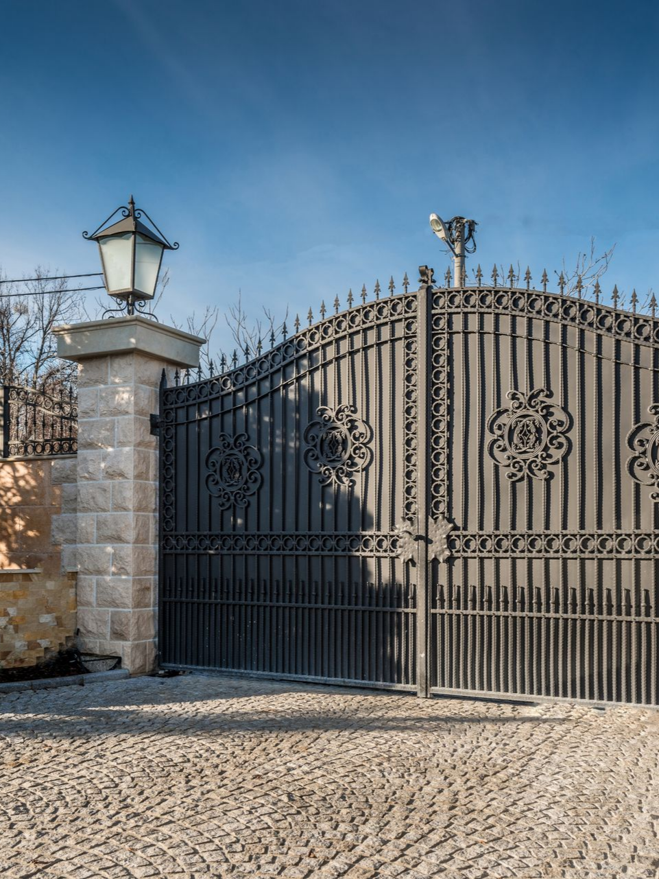 Portail en métal noir orné avec colonne en pierre et lanterne, sur une allée pavée sous un ciel bleu.