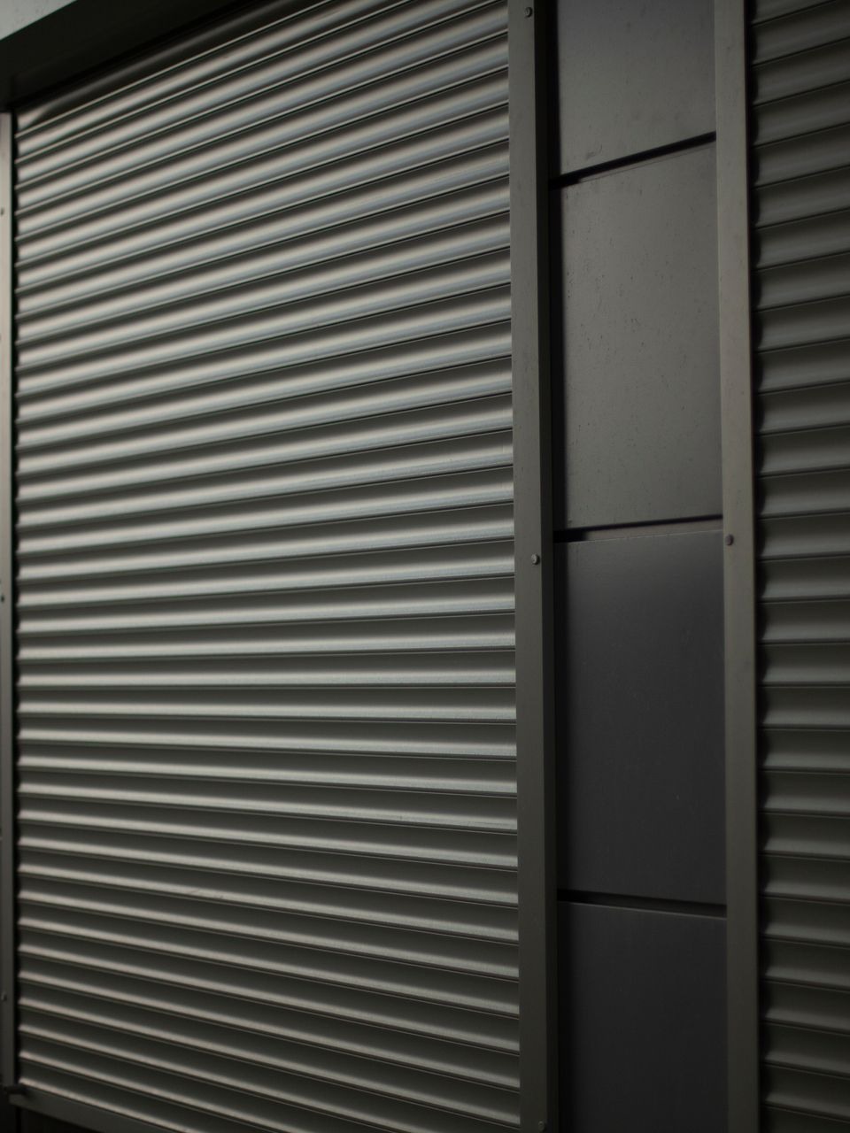 Volet roulant en métal gris avec lignes horizontales, adjacent à des panneaux gris foncé à l'extérieur du bâtiment.