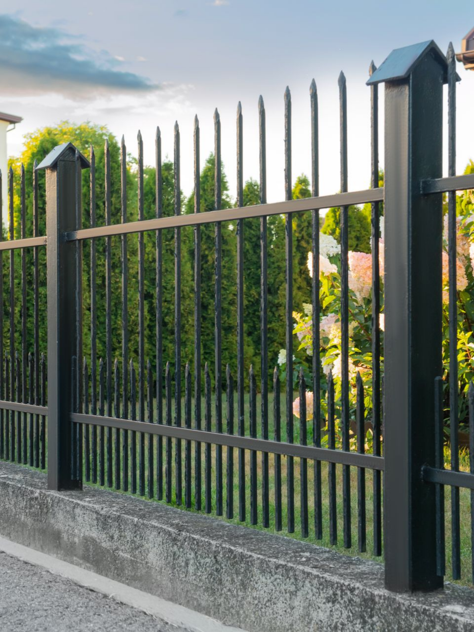 Clôture métallique noire à sommets pointus, posée sur un socle en béton, devant des arbres verts.