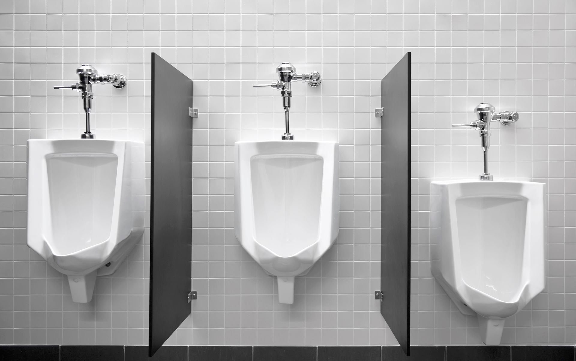 Trois urinoirs dans des toilettes publiques, séparés par des cloisons gris foncé, contre un mur carrelé.