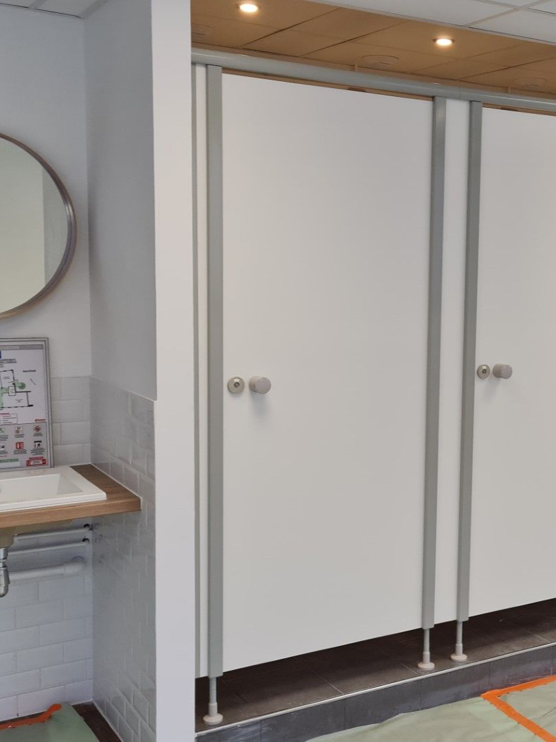 Cabines de salle de bains blanches avec quincaillerie argentée ; un lavabo et un miroir sont visibles sur la gauche.