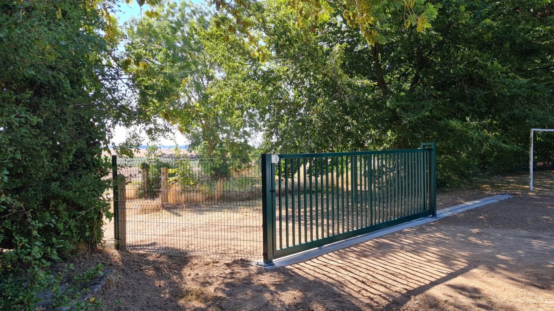 Portail métallique vert, ouvert, dans un chemin de terre menant à une zone mal entretenue avec des arbres.