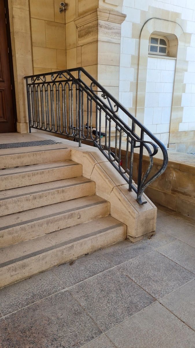 Escalier en pierre menant à une porte avec rampe en fer forgé noir. Pierre beige clair et sol gris.