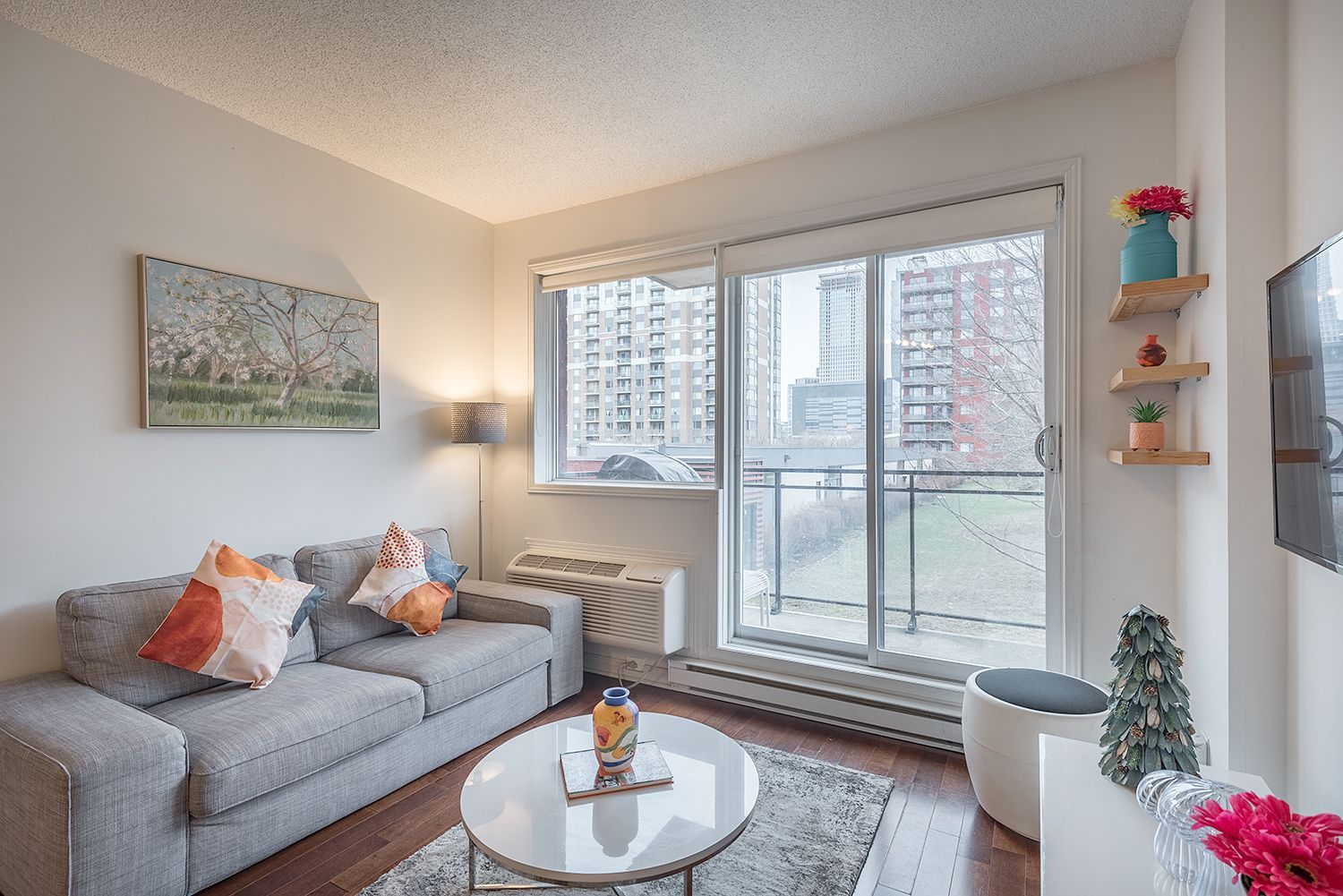 Un petit salon lumineux avec un canapé, une porte coulissante en verre et un balcon donnant sur les bâtiments de la ville.