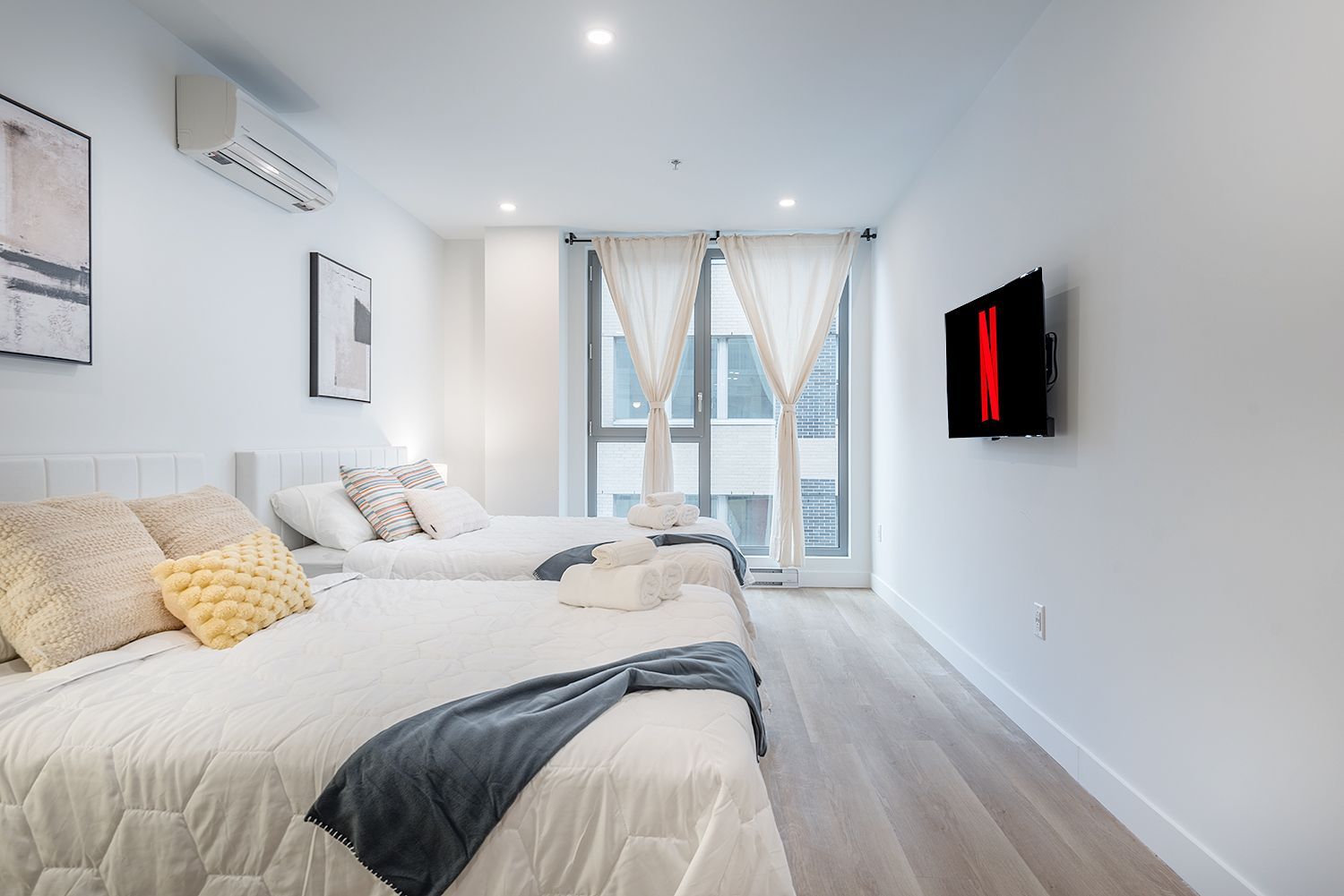 Chambre avec deux lits, murs blancs, une télévision et une grande fenêtre avec rideaux.