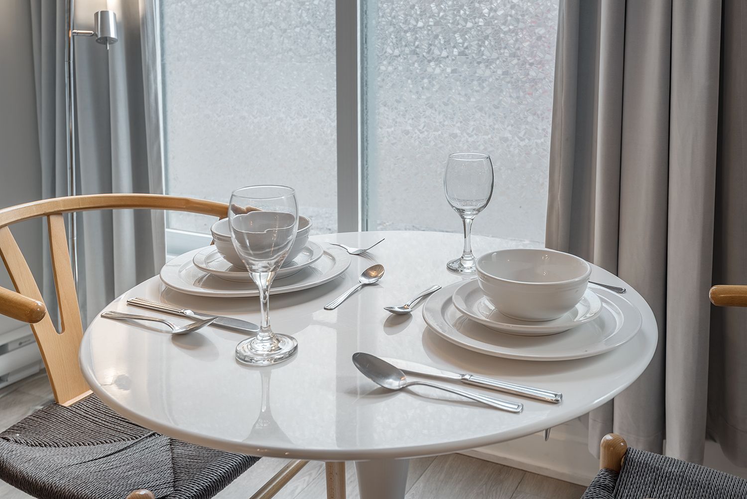 Une petite table à manger blanche pour deux personnes, près d'une fenêtre. Des chaises claires l'entourent.