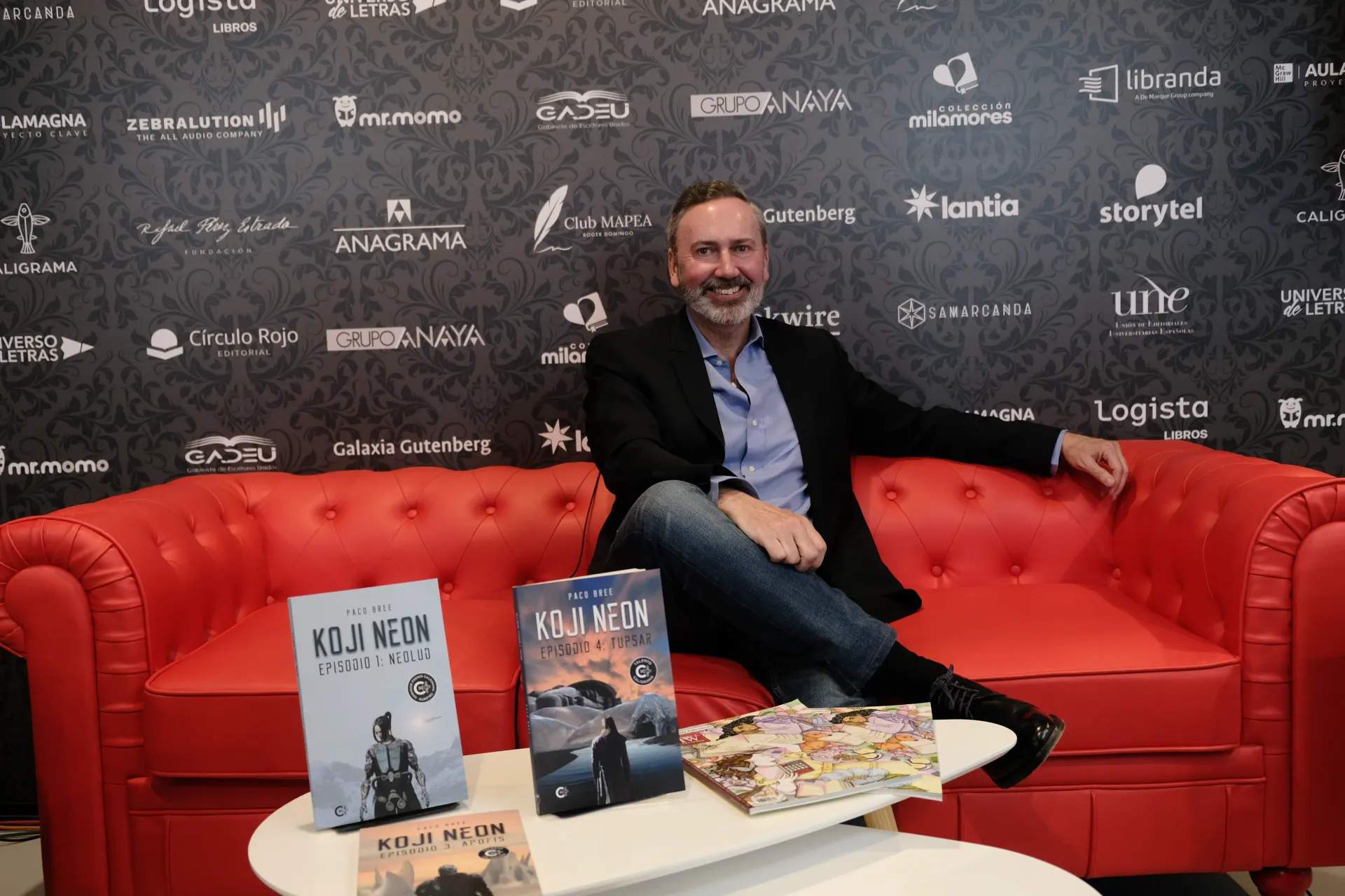 Un hombre está sentado en un sofá rojo al lado de una mesa con libros encima.