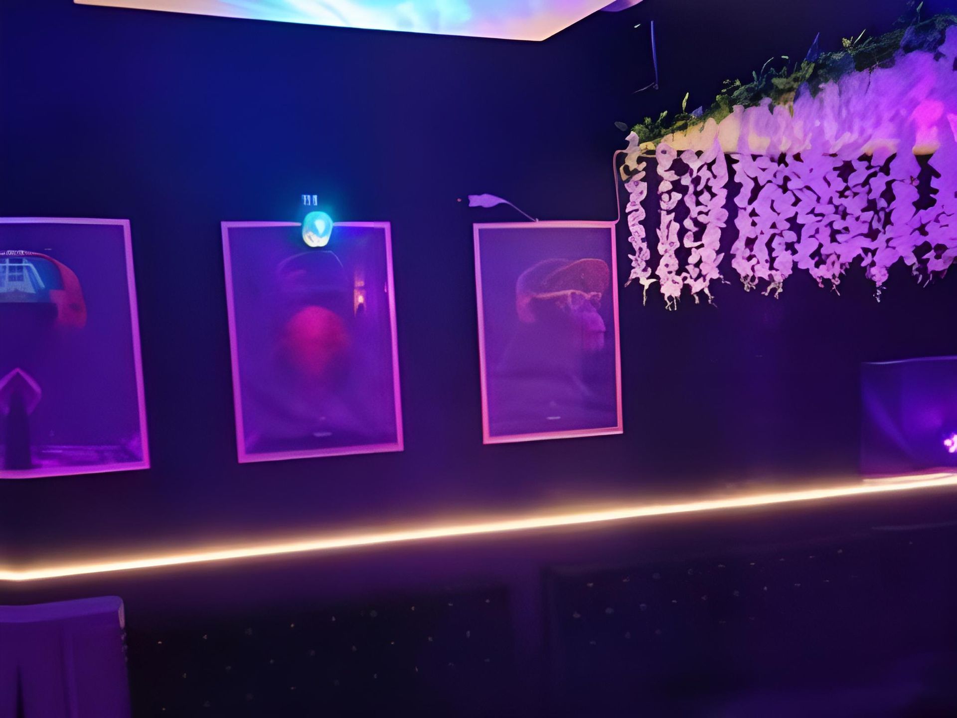 Mur noir orné de trois œuvres encadrées, éclairées par des lumières violet et bleu. Un bandeau lumineux au néon court en dessous.