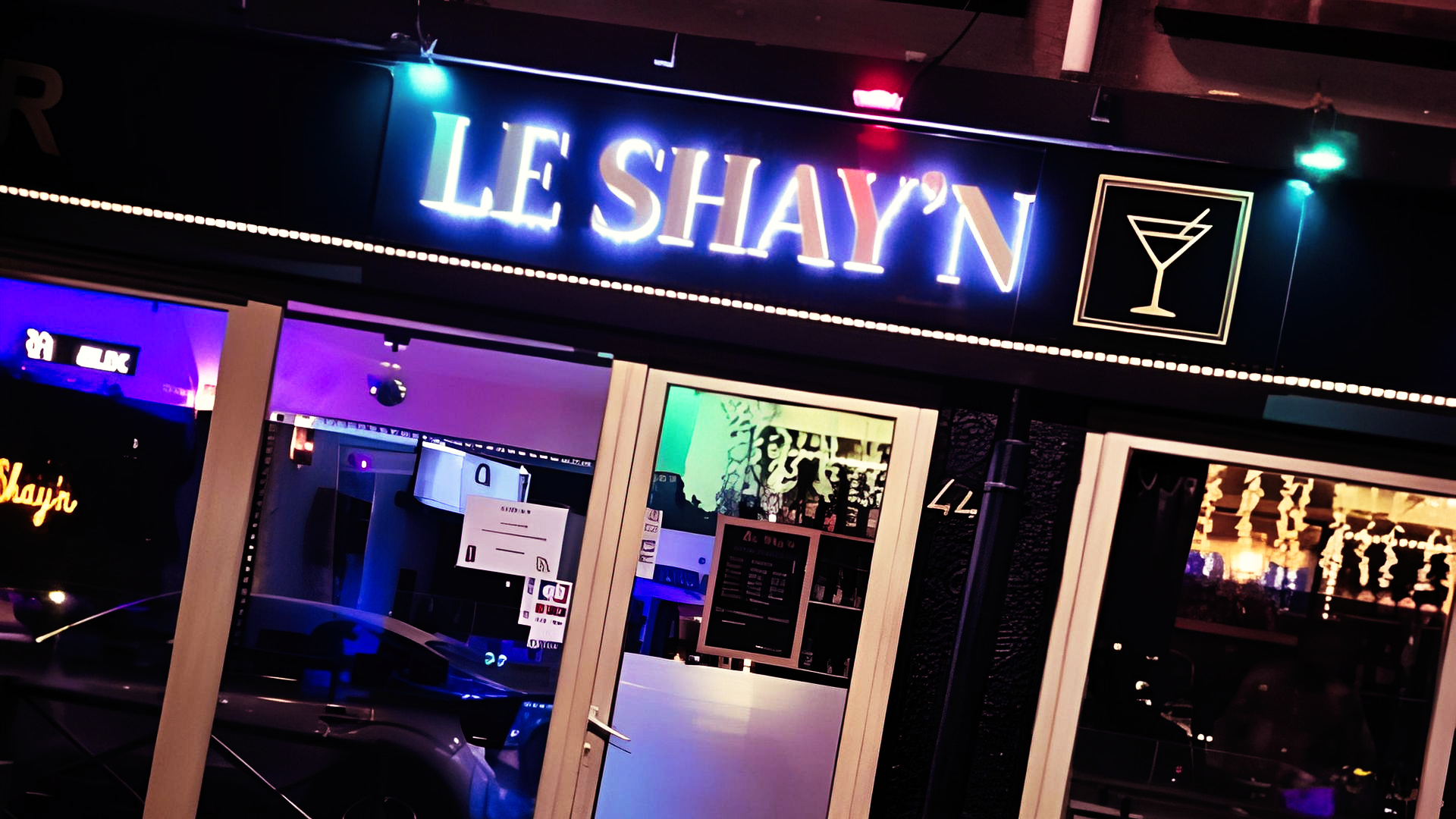L'entrée du bar, ornée d'un verre à cocktail et d'une enseigne lumineuse. Intérieur visible, éclairé de lumières colorées.