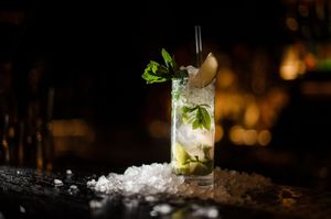 Un mojito servi dans un grand verre, garni de menthe et de citron vert, entouré de glace sur un bar sombre.