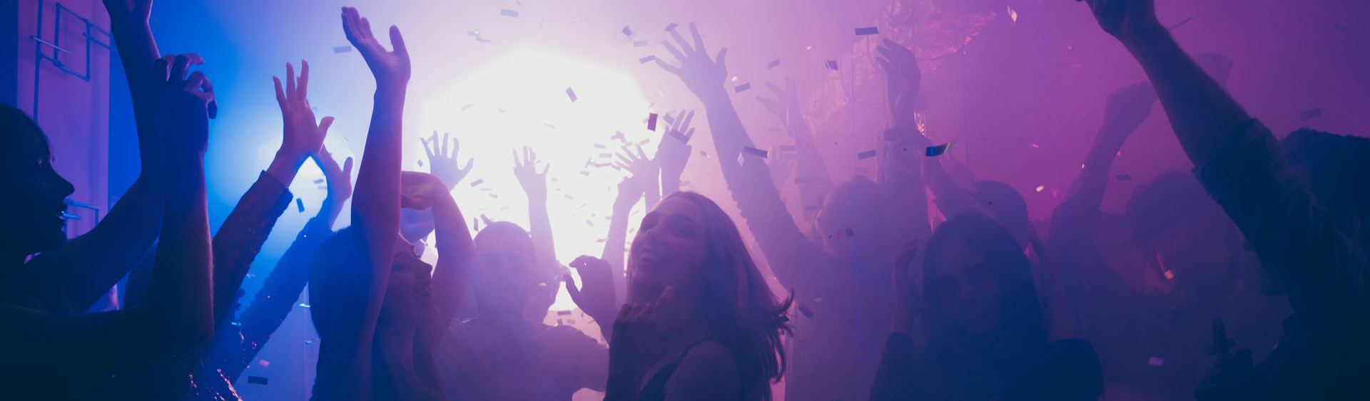 Des gens dansent les bras levés dans une boîte de nuit sous des lumières violet et bleu, des confettis volent.