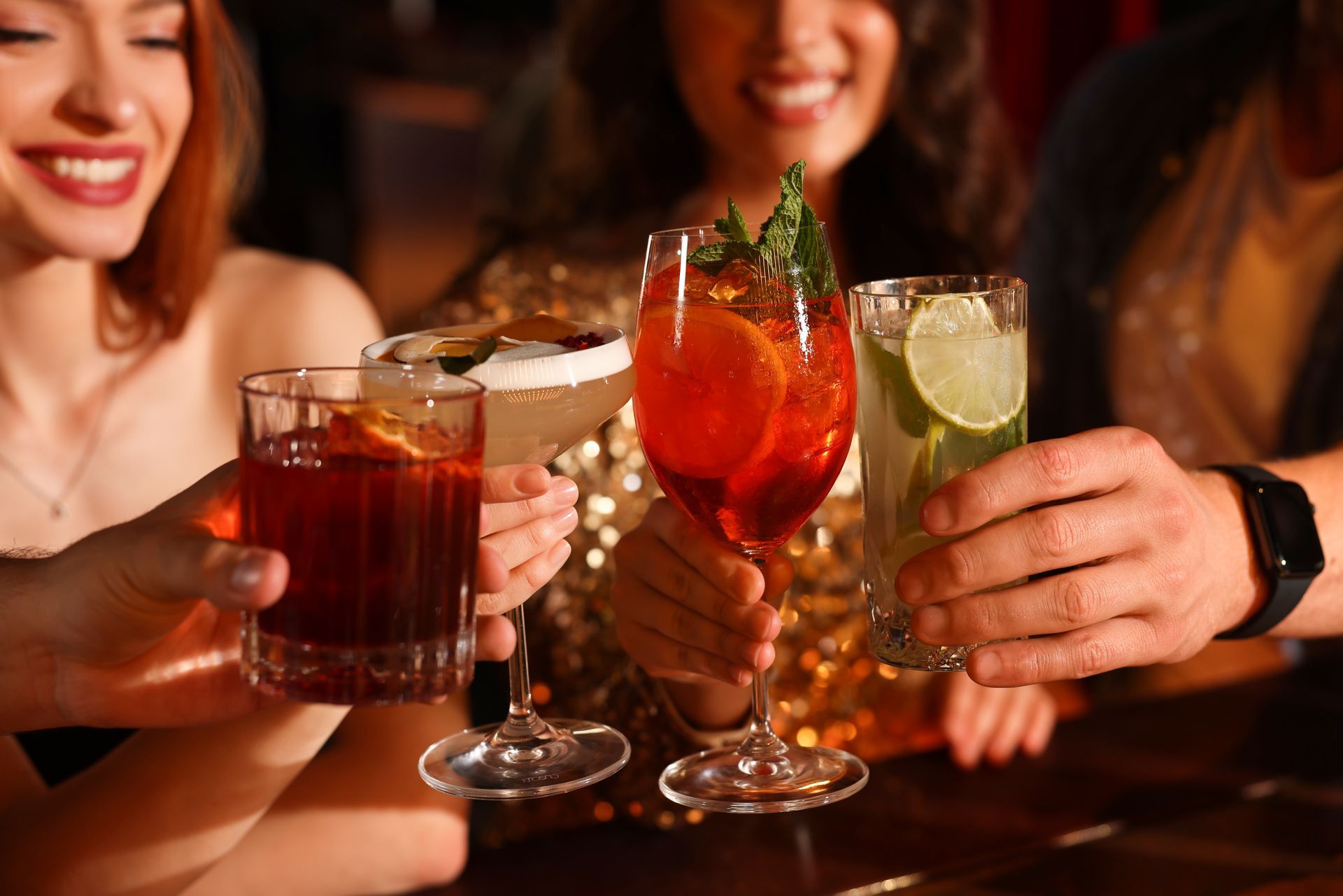 Des personnes trinquent avec des cocktails dans un bar. Les cocktails sont orange, vert et rouge.