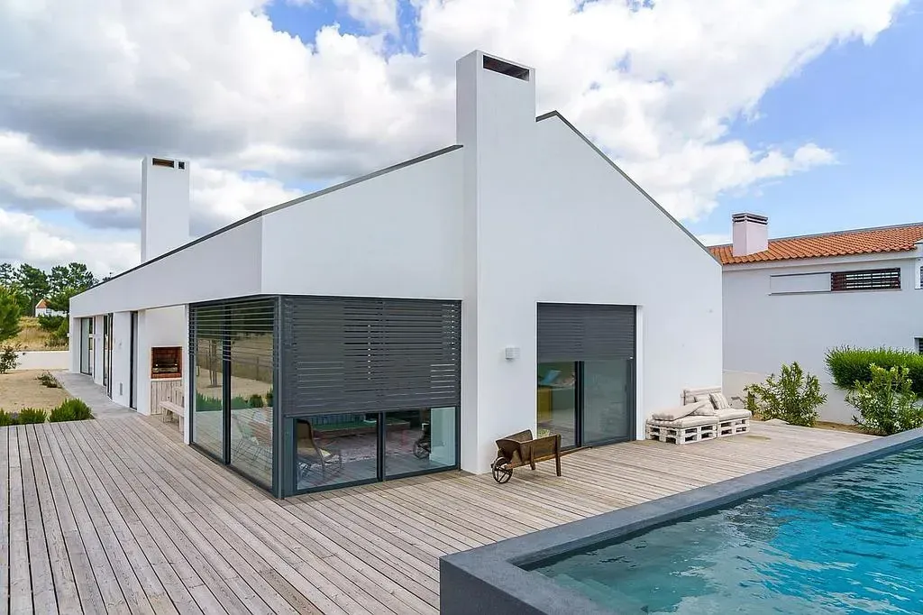 Maison moderne blanche avec piscine, terrasse en bois et murs en verre, sous un ciel nuageux.