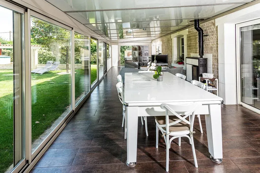 Longue table à manger dans la véranda avec murs en verre, vue sur la pelouse, chaises blanches et plafond.