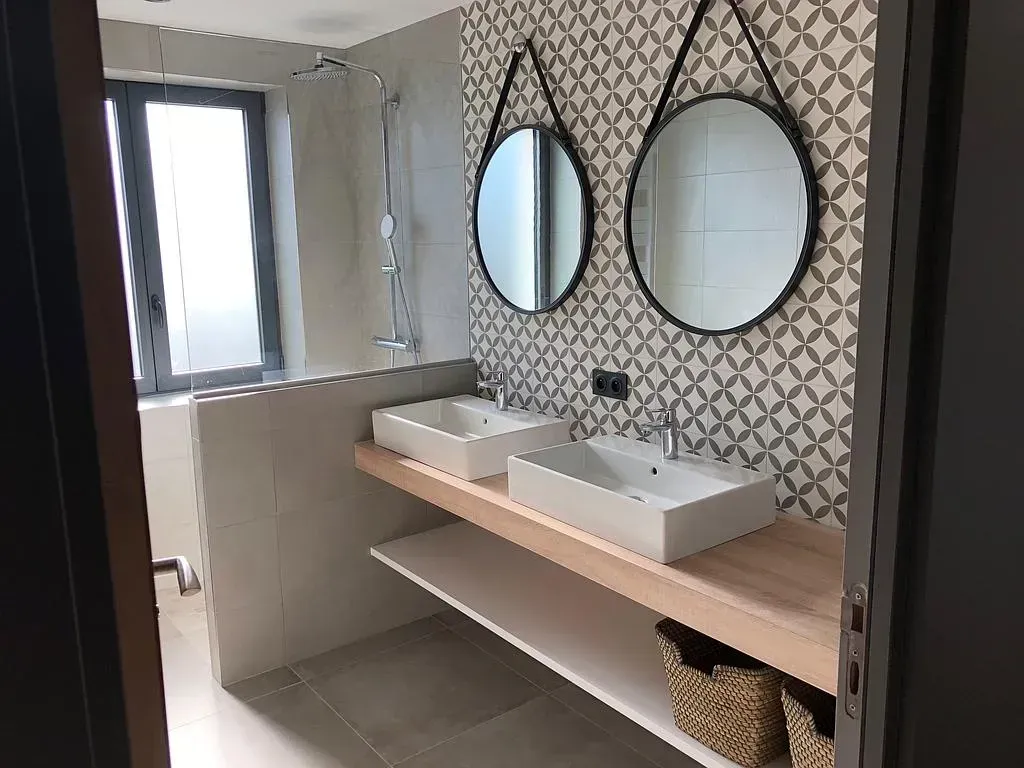 Salle de bains avec deux vasques, miroirs et papier peint à motifs. Lumière naturelle et touches de bois.