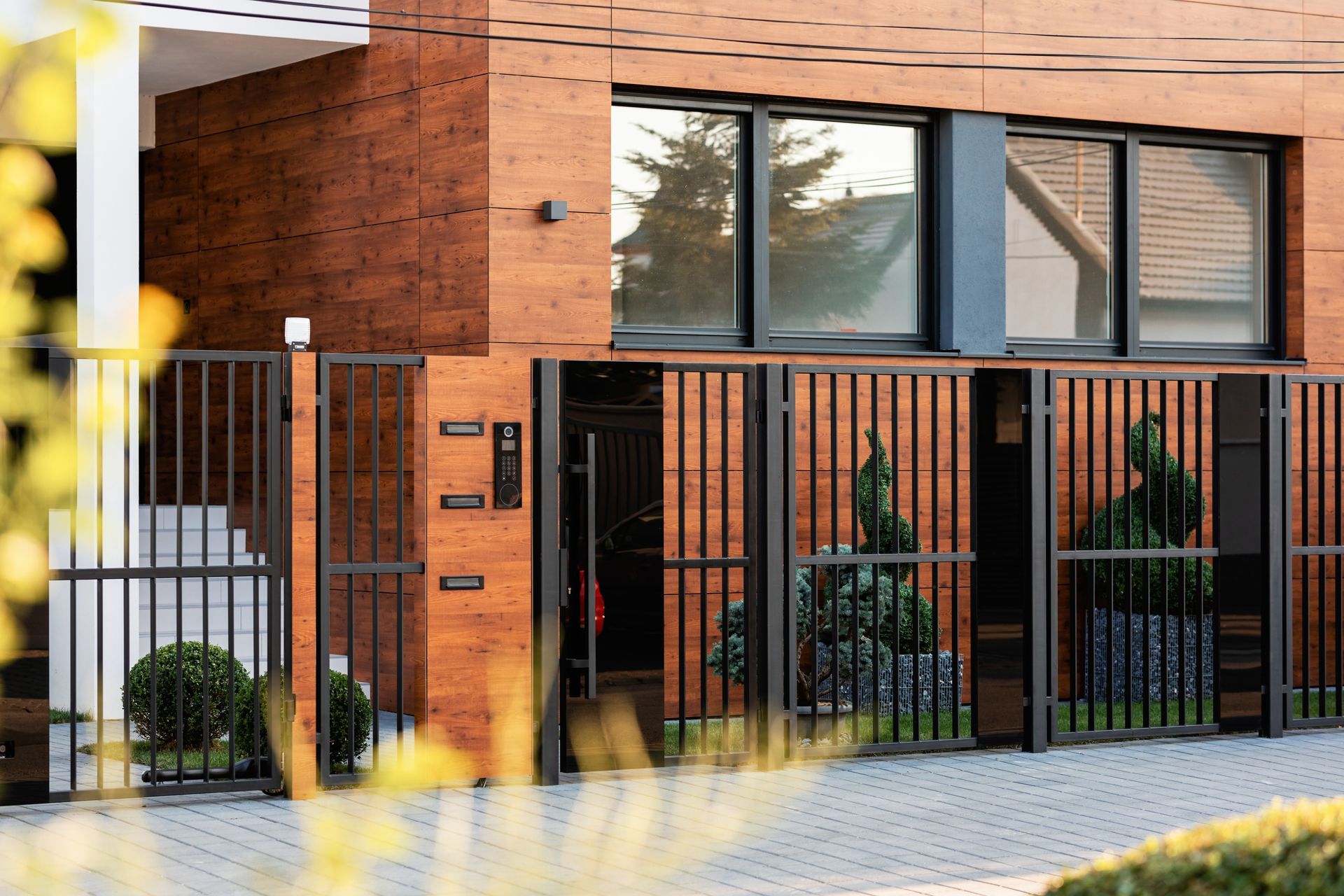 Extérieur de maison moderne avec façade en bois, clôture en métal noir et aménagement paysager soigné.