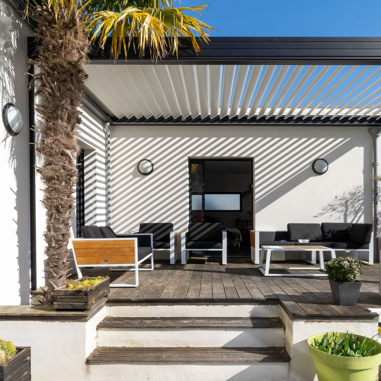 Patio avec pergola, palmier et mobilier moderne projetant des ombres rayées sur la maison blanche.