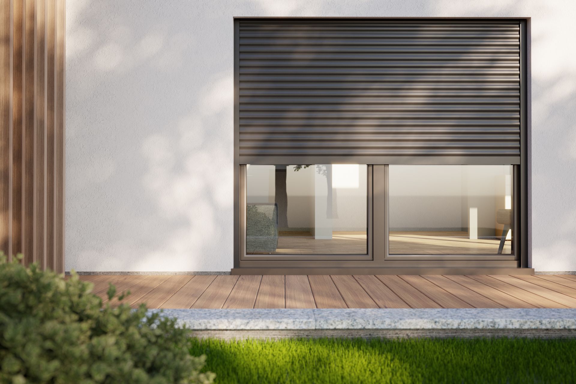 Fenêtre avec volet roulant gris foncé fermé sur un bâtiment blanc, sur une terrasse en bois avec une pelouse verte.