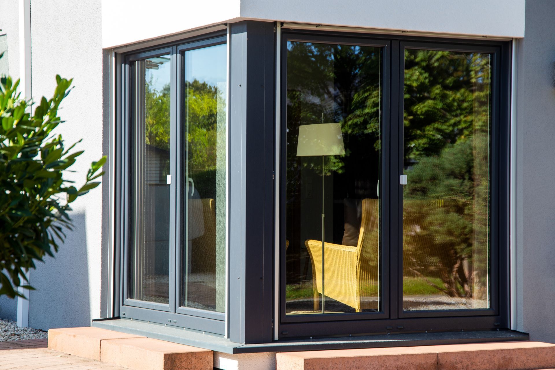 Véranda aux parois de verre avec cadre gris foncé, laissant entrevoir une chaise jaune et une lampe à l'intérieur.