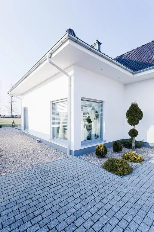 Maison blanche avec allée pavée et petit jardin avec buissons.