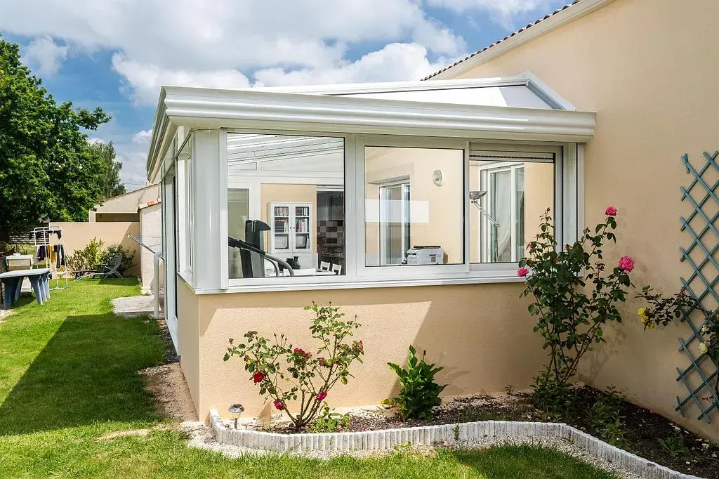 Ajout d'une véranda avec cadre blanc, fenêtres et un muret beige, attaché à une maison beige.