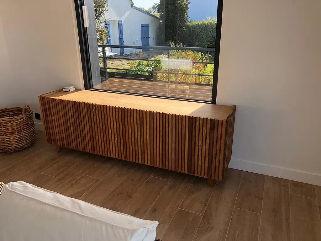Armoire en bois avec façade texturée, placée sous une fenêtre, à côté d'un panier en osier, dans une pièce avec parquet.