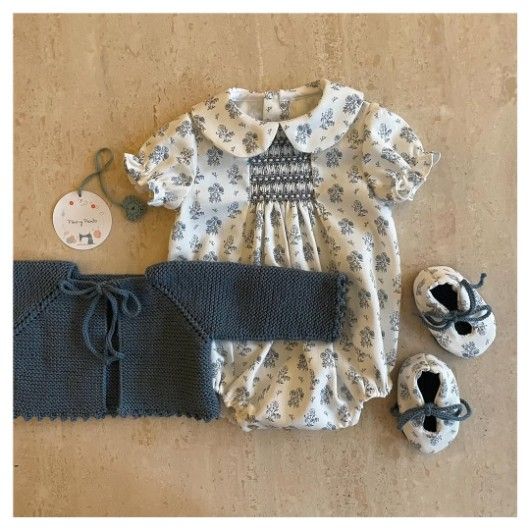 Conjunto de bebé: mono floral, suéter azul y patuquitos a juego sobre superficie color canela.