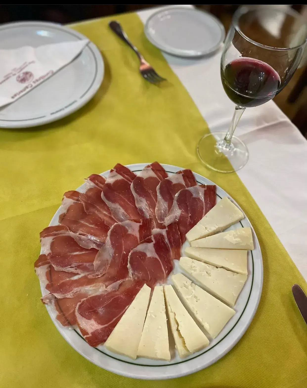 Plato de jamón y queso, con una copa de vino tinto. Mantel amarillo y blanco.
