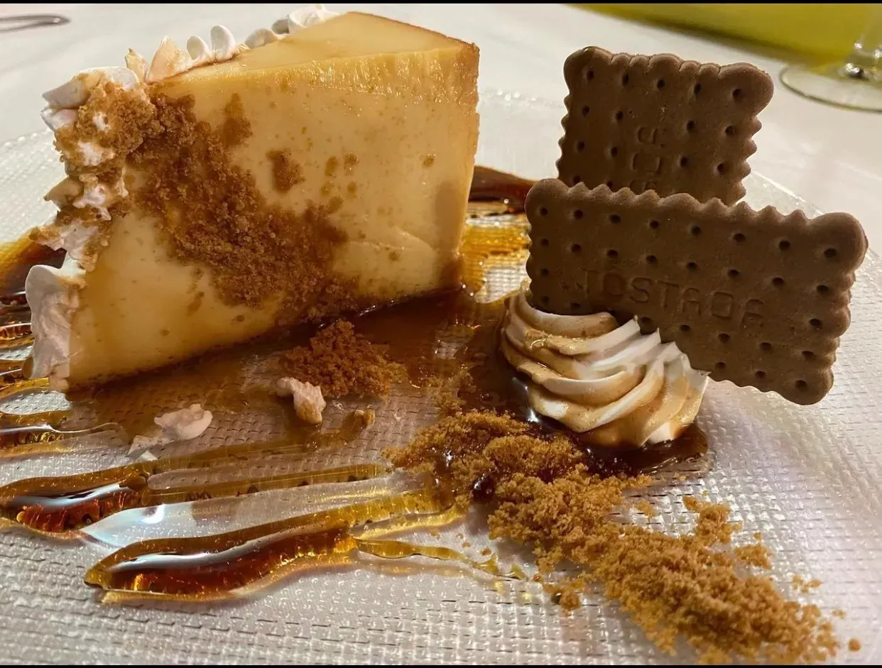 Postre de flan con salsa de caramelo, crema batida, migas trituradas y galletas en un plato transparente.