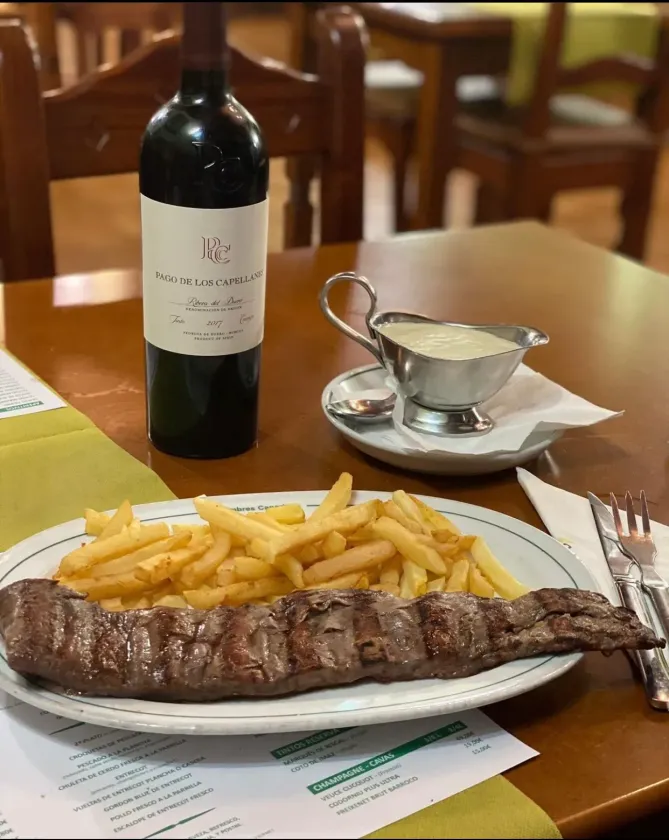 Bistec con patatas fritas, botella de vino y salsera en una mesa de restaurante.