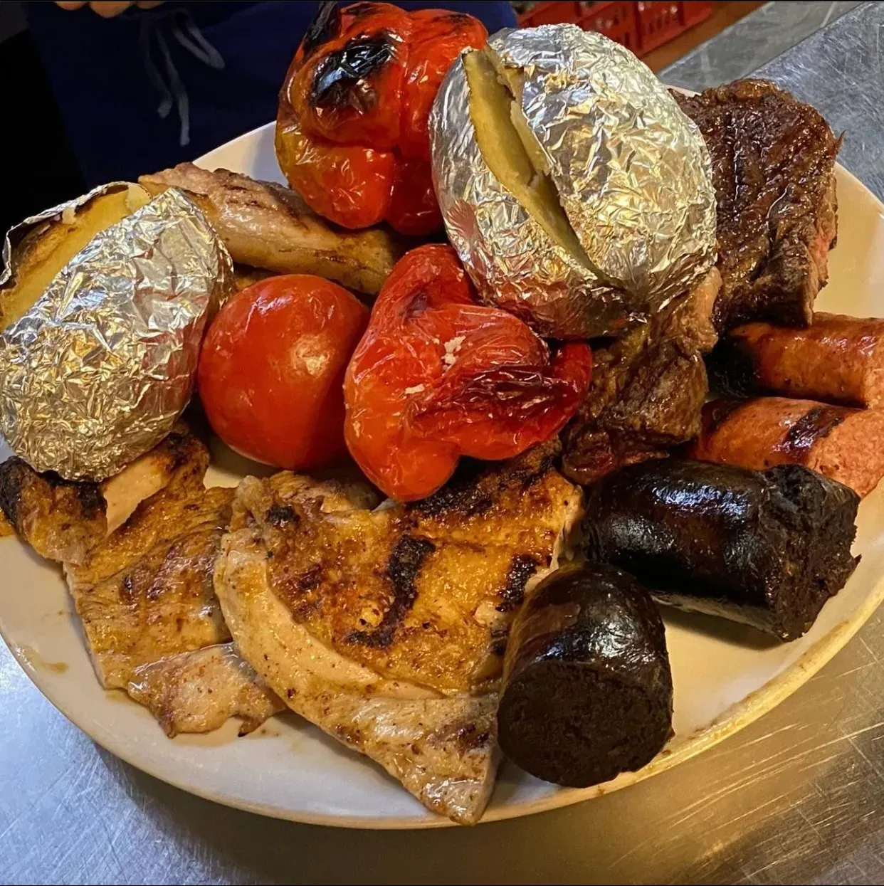Carnes a la parrilla, verduras y patatas al horno en un plato, incluyendo pimientos rojos, tomate y salchichas.