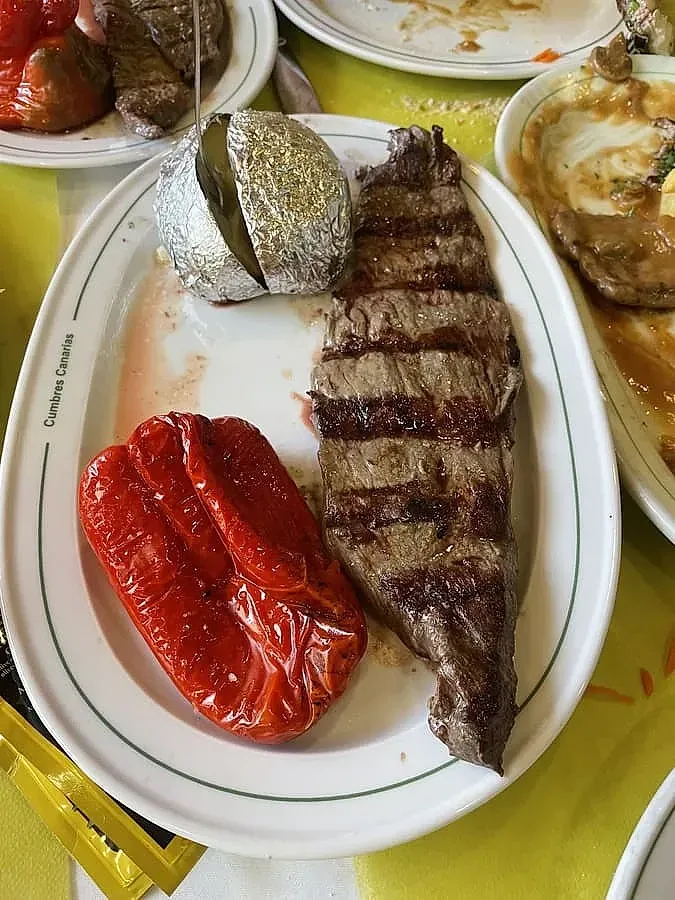 Bistec a la parrilla, pimiento rojo, papa al horno en el plato; otros platos visibles.