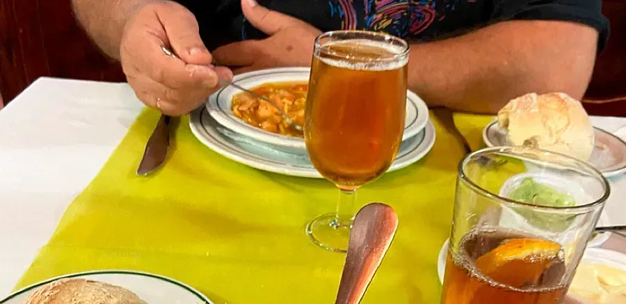 Persona comiendo sopa y bebiendo cerveza. Mesa puesta con pan, mantequilla y vaso.