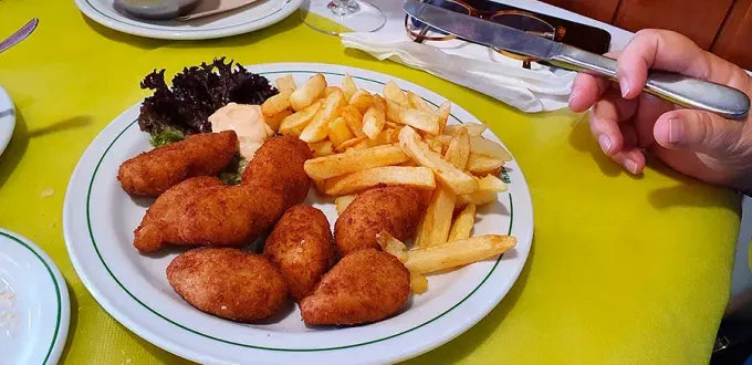 Plato de comida frita con patatas fritas, croquetas, lechuga y salsa sobre una mesa amarilla, sostenido por una mano con un cuchillo.