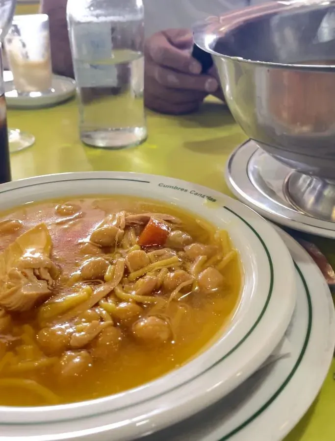 Cuenco de sopa con garbanzos y fideos sobre una mesa con una persona al fondo.