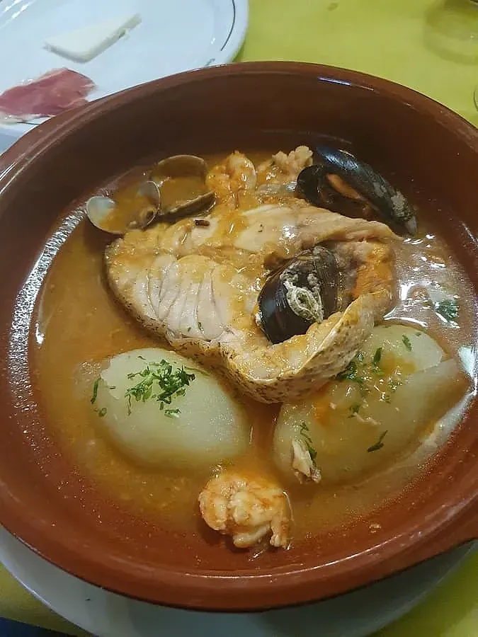 Guiso de salmón en recipiente marrón con patatas, camarones, mejillones y caldo; plato blanco con queso y carne de fondo.