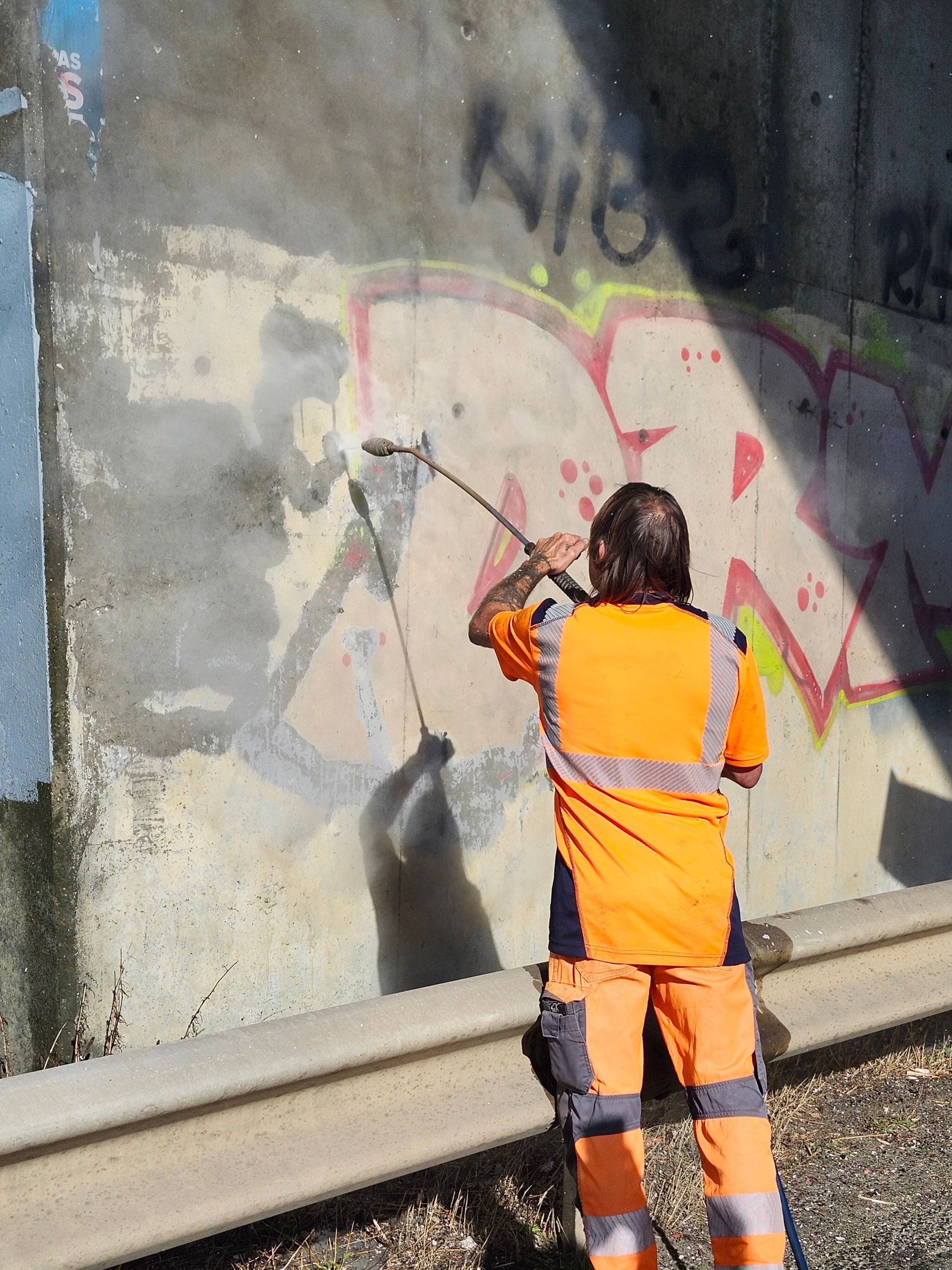 Une personne portant un gilet de sécurité orange nettoie à haute pression des graffitis sur un mur en béton extérieur.