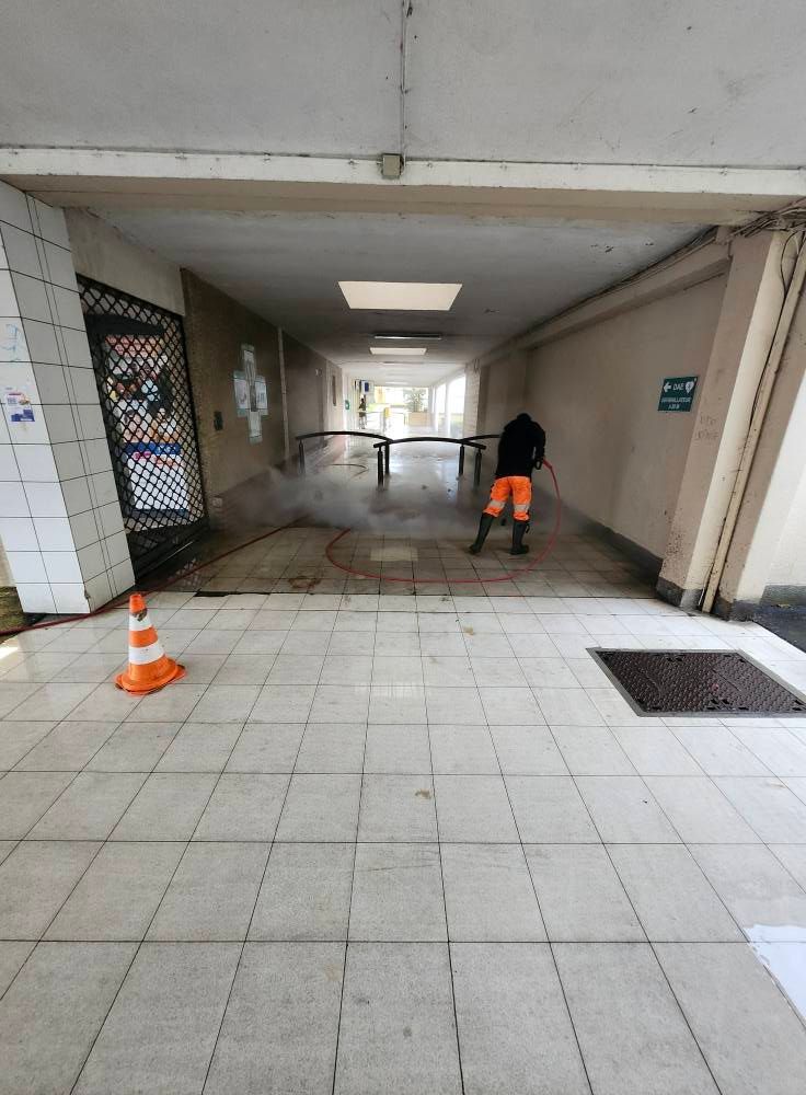 Une personne nettoie un couloir carrelé au nettoyeur haute pression ; un cône orange et un panneau de sortie sont visibles.