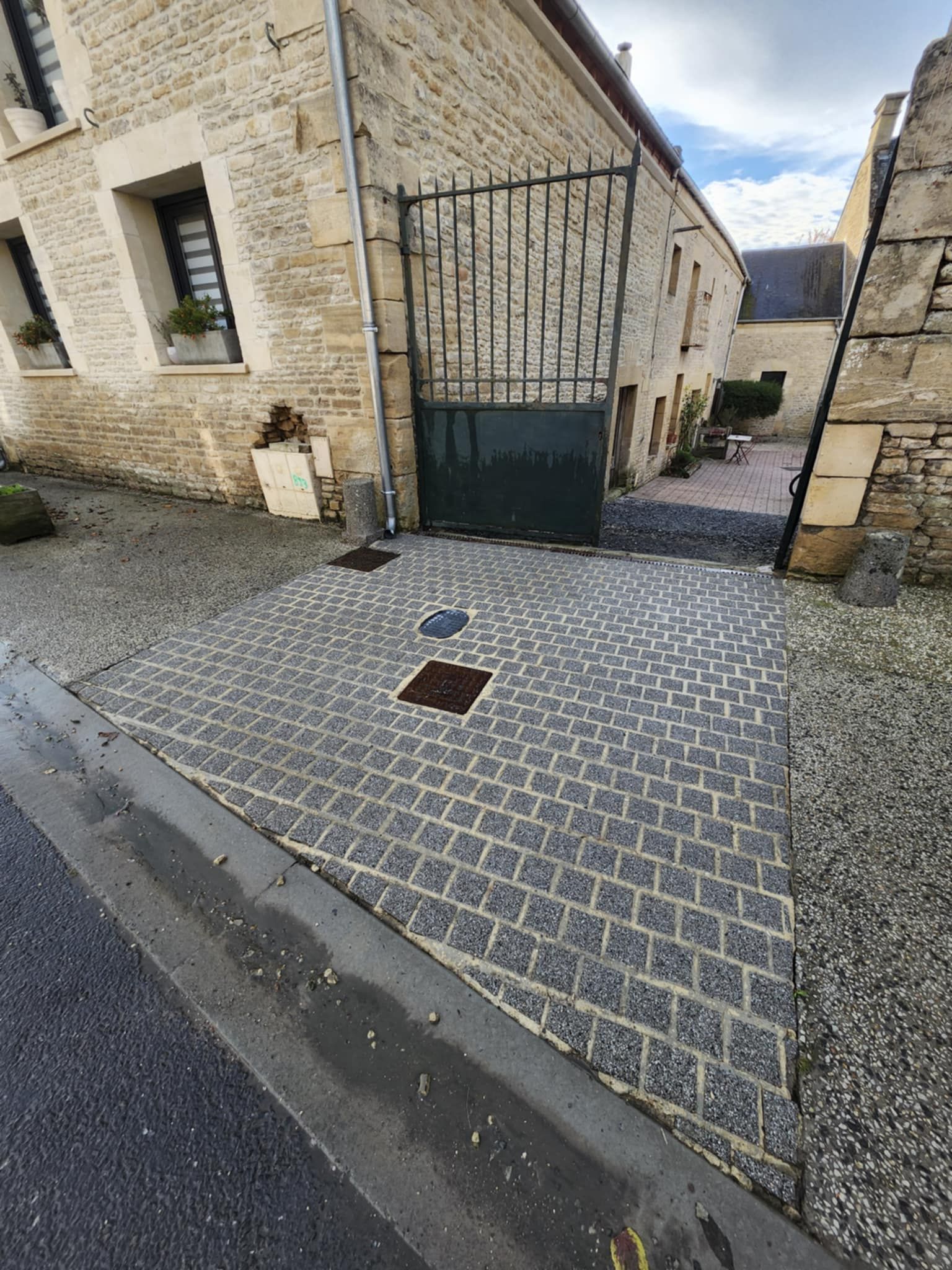 Allée pavée menant à une entrée fermée par un portail, entre des bâtiments en pierre.