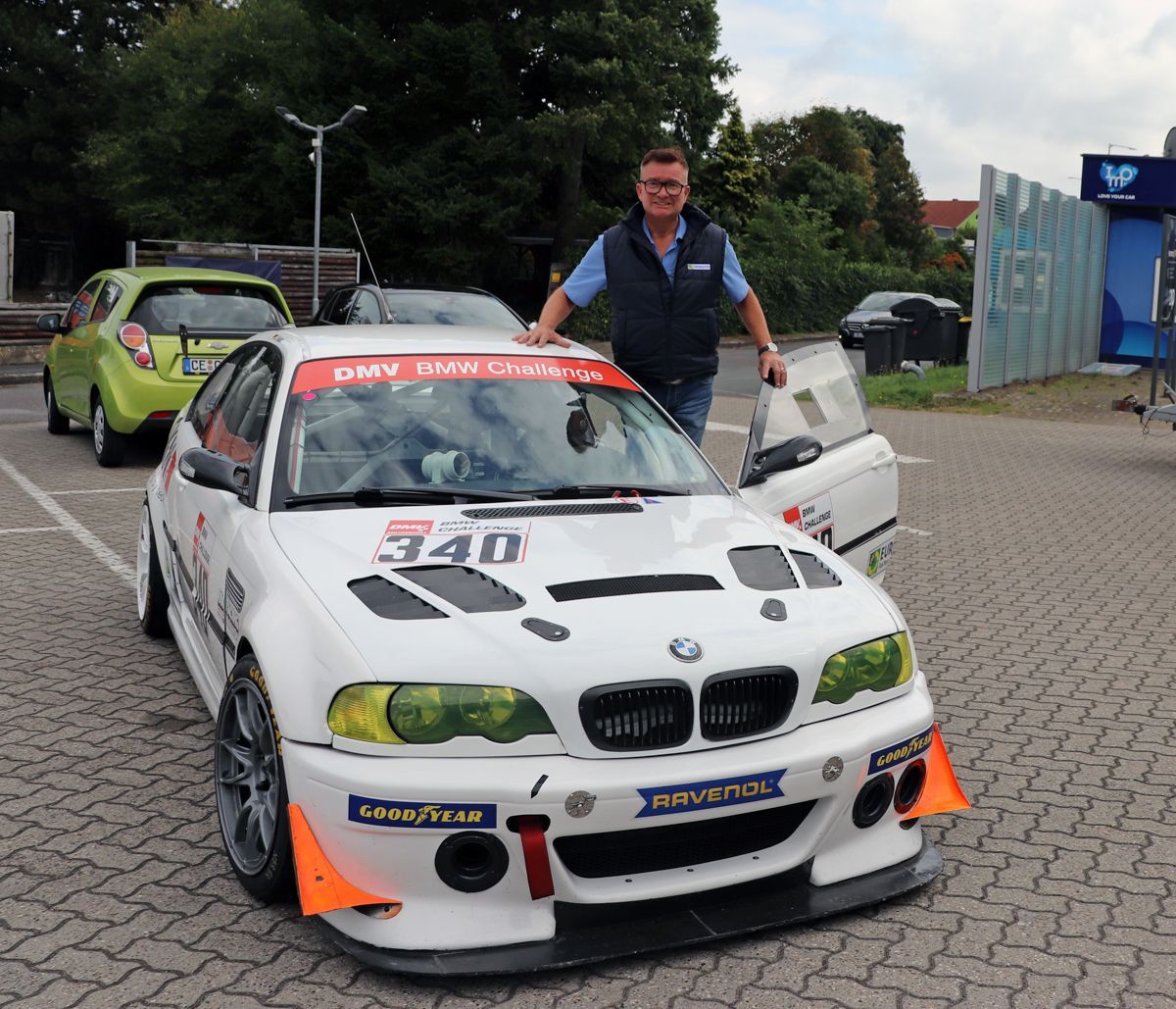 Ein Mann steht neben einem weißen BMW-Rennwagen, der im Freien geparkt ist.