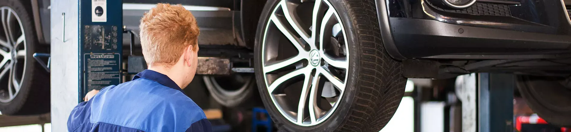 Ein Mechaniker arbeitet an einem auf einer Hebebühne angehobenen Auto. Der Mechaniker trägt eine blaue Arbeitskleidung.