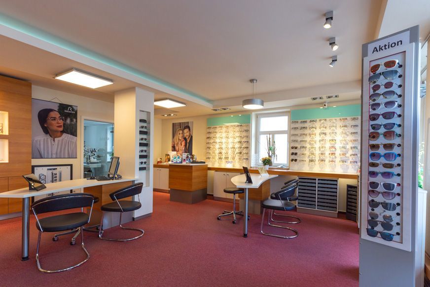 Innenraum einer Optikerpraxis mit Brillendisplays, Stühlen und Theken. Roter Teppich, helle Beleuchtung.