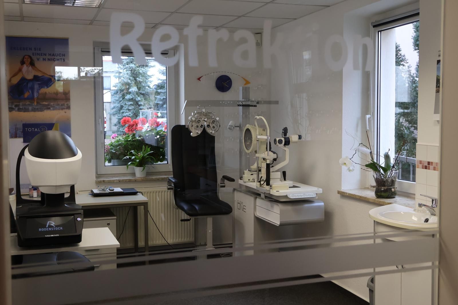 Innenansicht einer Optikerpraxis mit Geräten, Stuhl und der Aufschrift „Refrakton“ auf der Glastür.