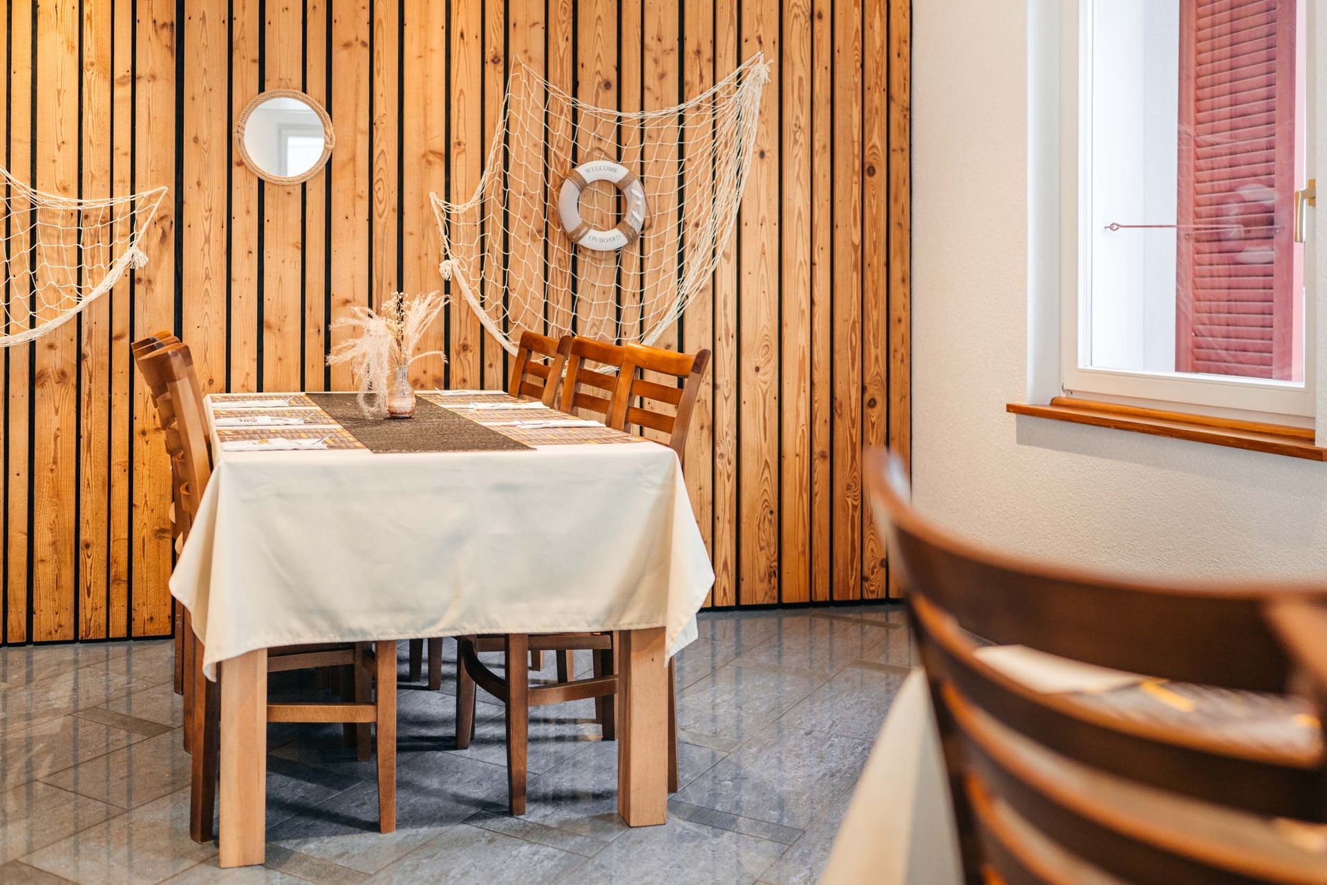 Essbereich im maritimen Stil: Holzwände, gedeckter Tisch für Gäste, Bullauge, Netz, Rettungsring.